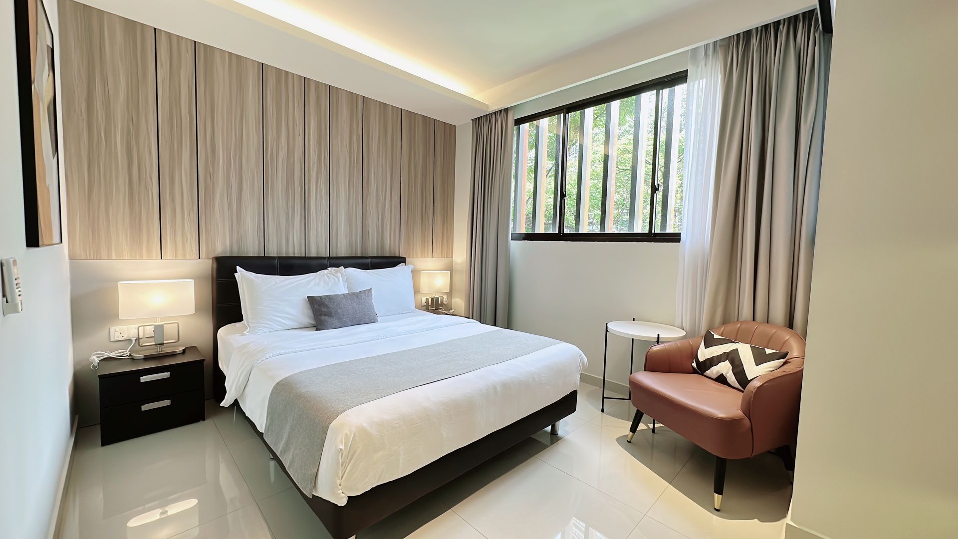 Modern bedroom with queen bed, bedside lamp, large window, and tan armchair