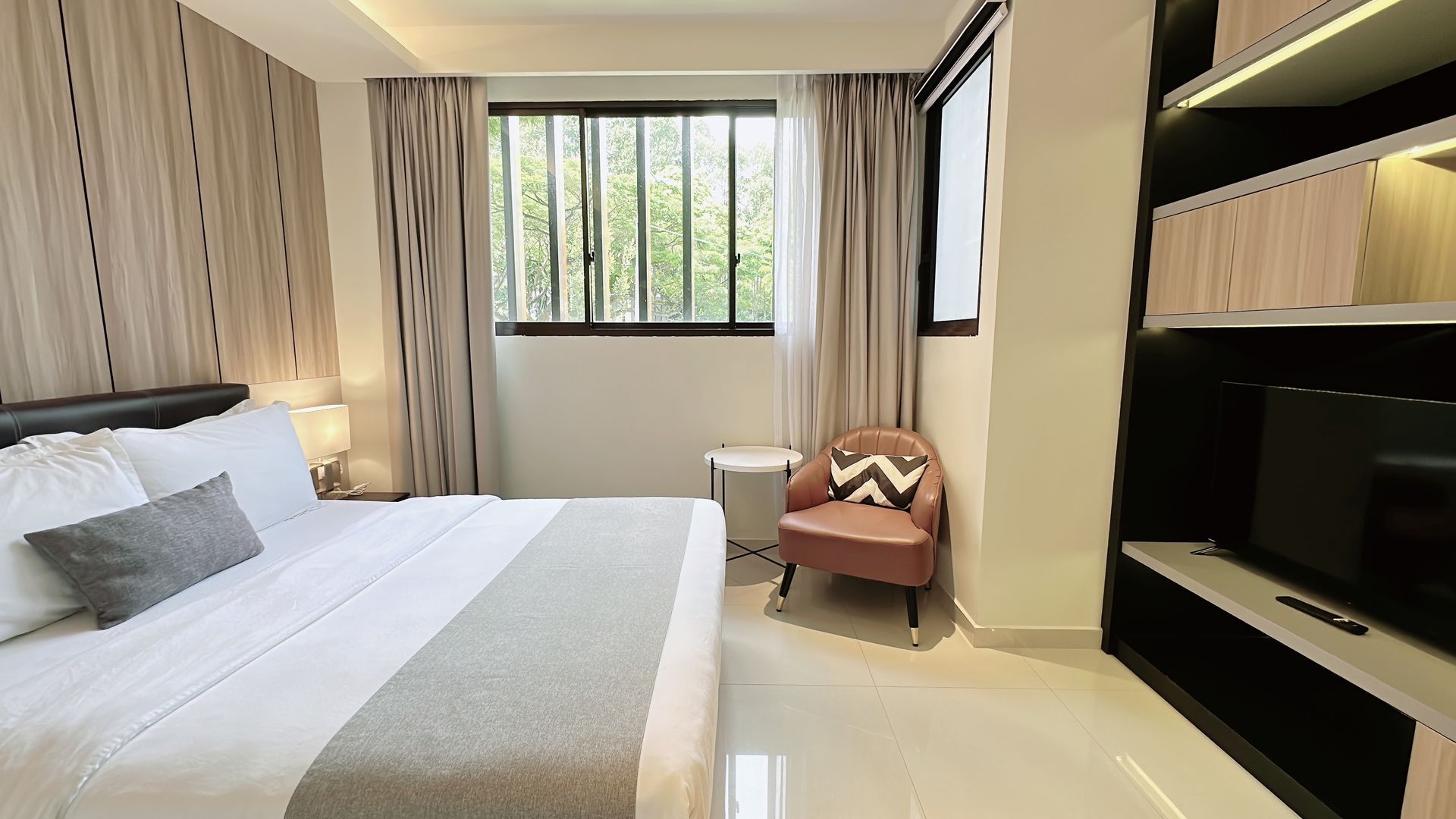 Modern bedroom with bed, window, armchair, and wall-mounted TV in a bright, minimalist room