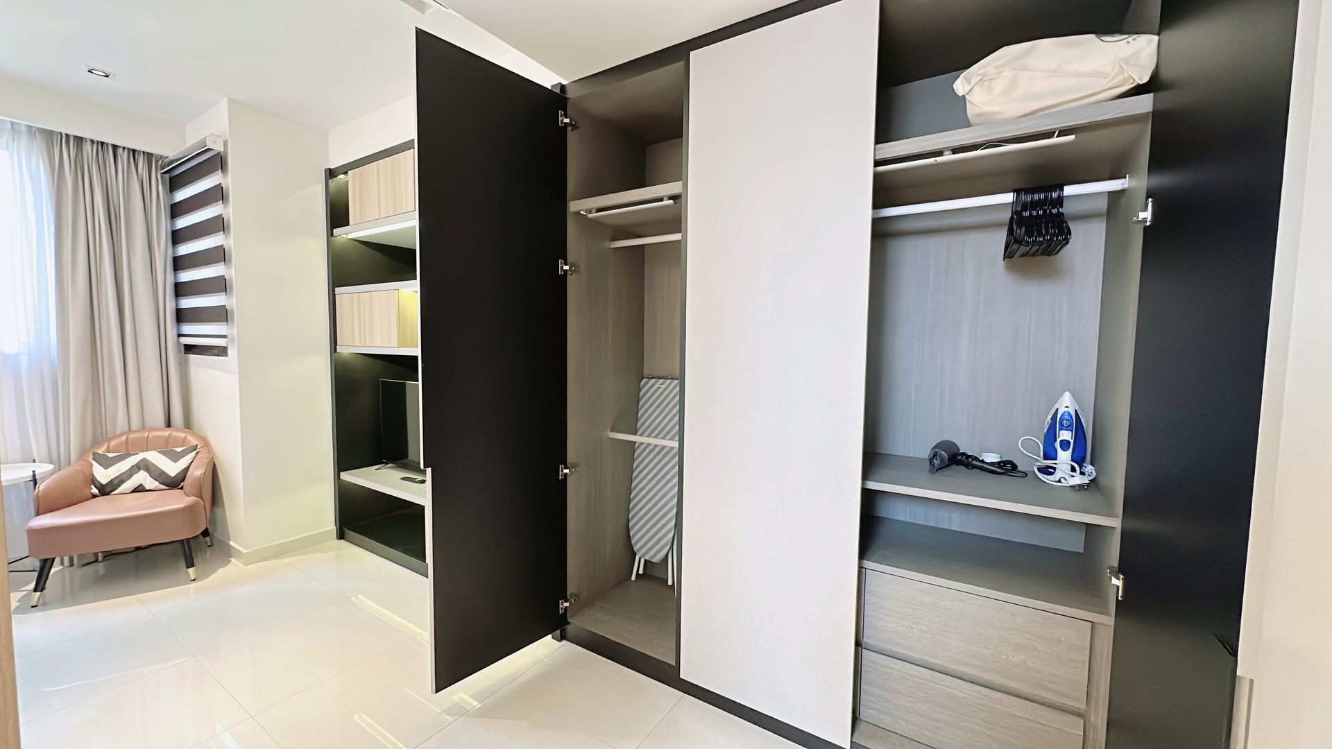 Open wardrobe with black, white, and gray doors in a bright bedroom; chair and curtains on the left.