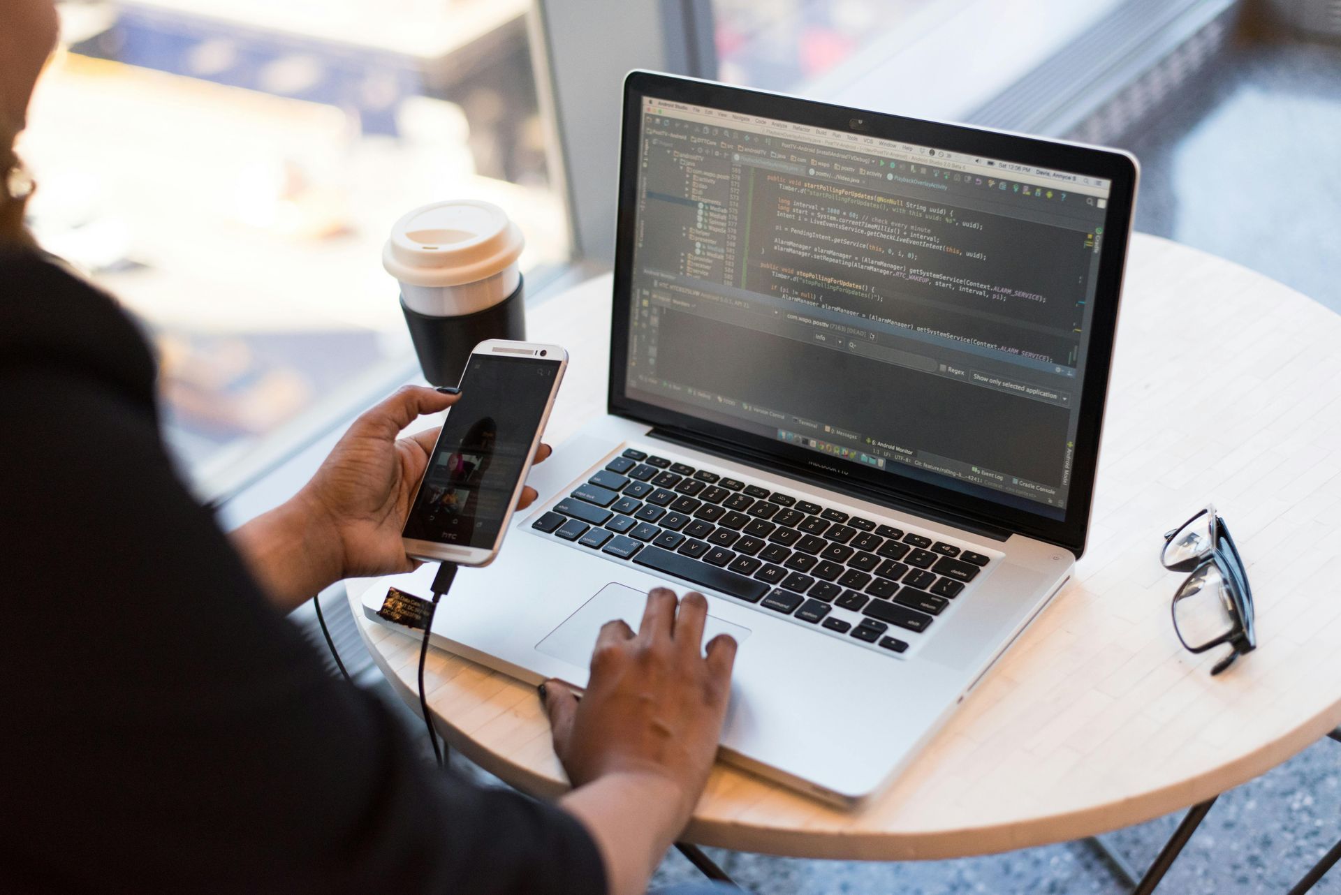 Person coding on a laptop, holding a phone, with a coffee cup and glasses on a table.
