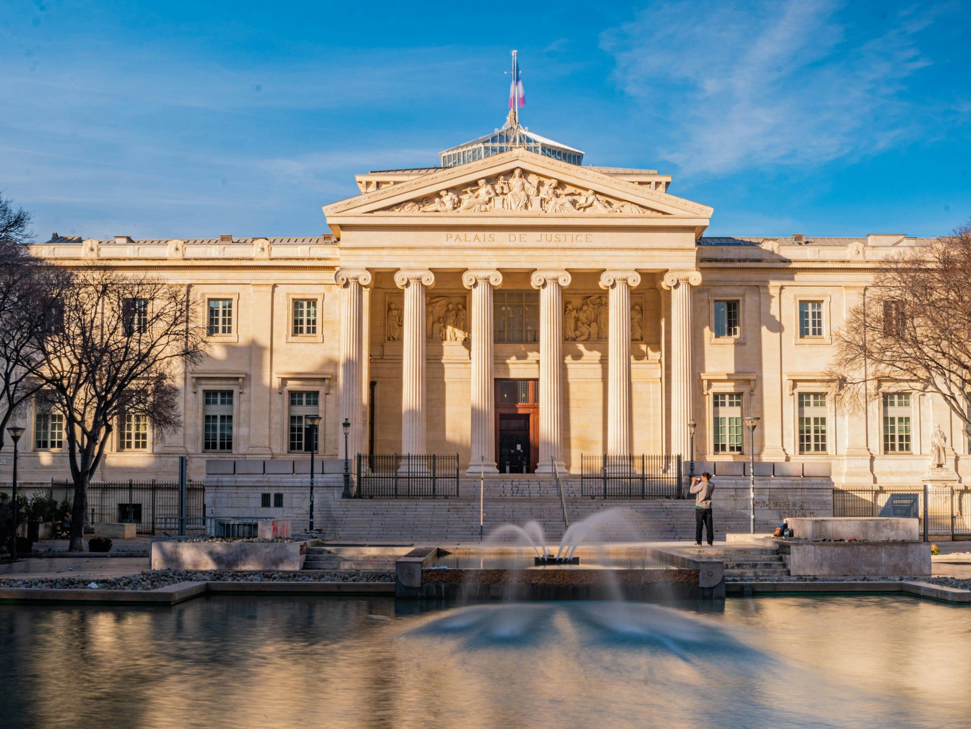 Palais de justice avec colonnes, fontaine et ciel bleu. Une personne se tient près de la fontaine.