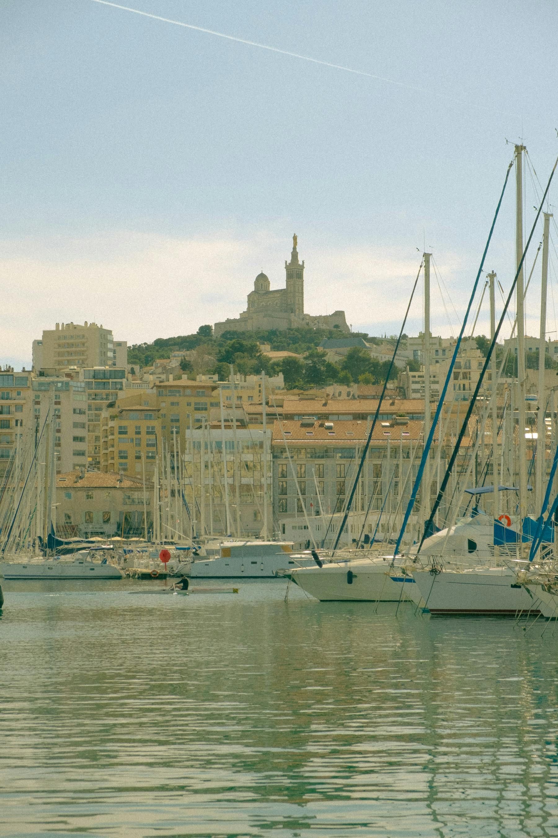 Voiliers dans un port, paysage urbain avec bâtiments et basilique sur une colline à Marseille, France.