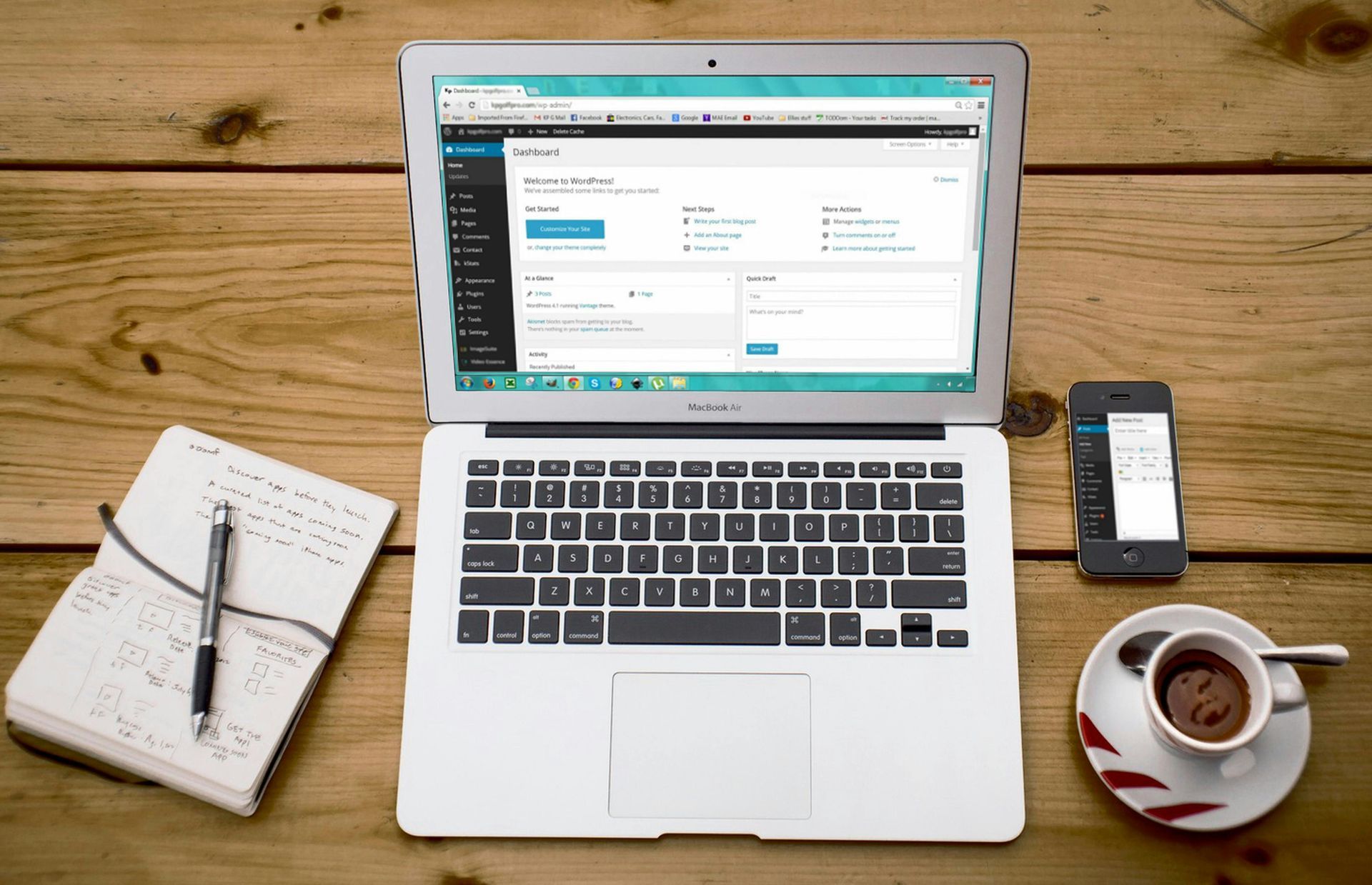 Laptop, phone, coffee, notebook, and pen on a wooden desk, open laptop displaying a web interface.