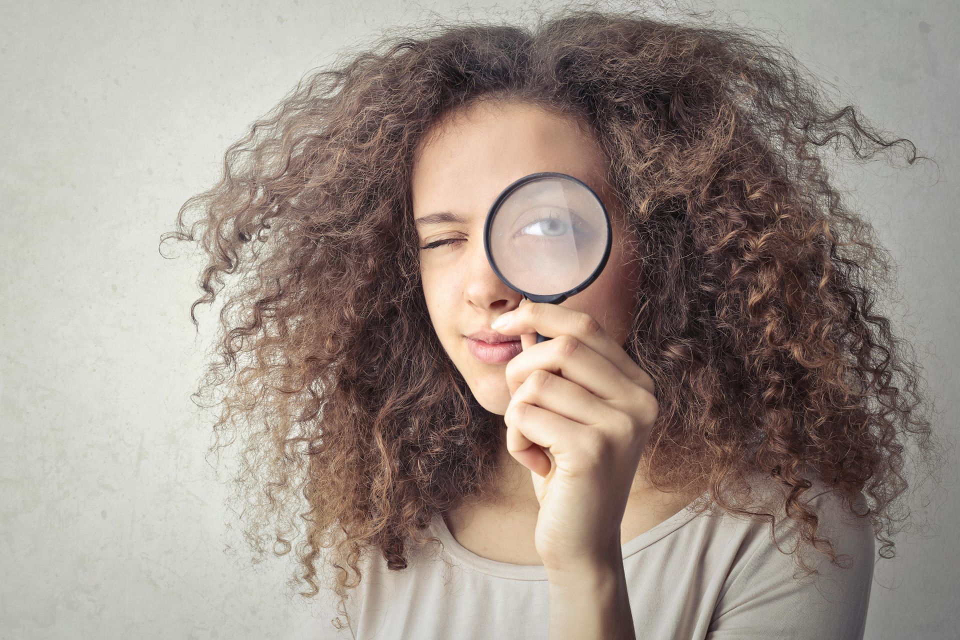 Woman with curly hair looking through a magnifying glass, squinting, against a neutral background.