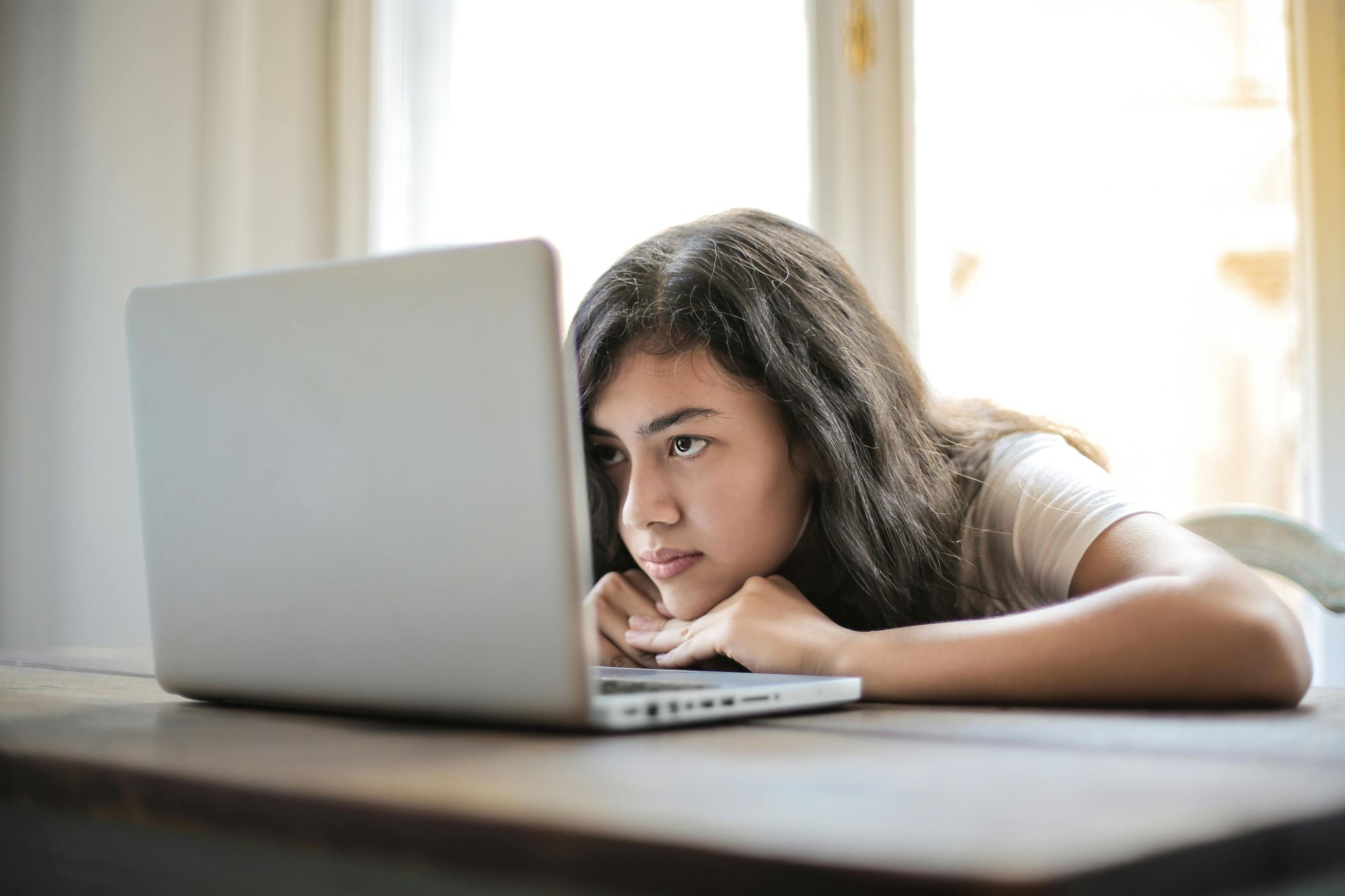Woman with long dark hair, resting chin on hands, looking intently at laptop screen at a table.