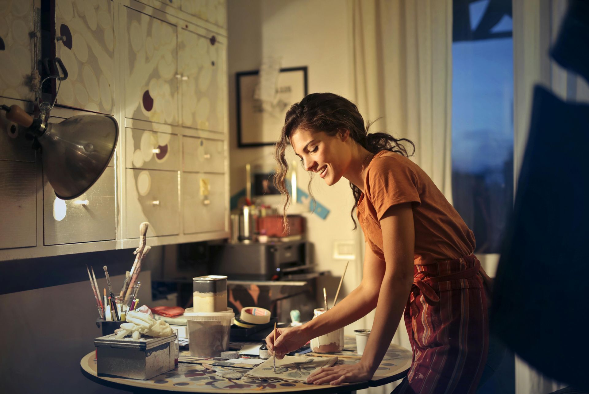 Woman smiling while painting at a desk in an art studio, warm lighting.