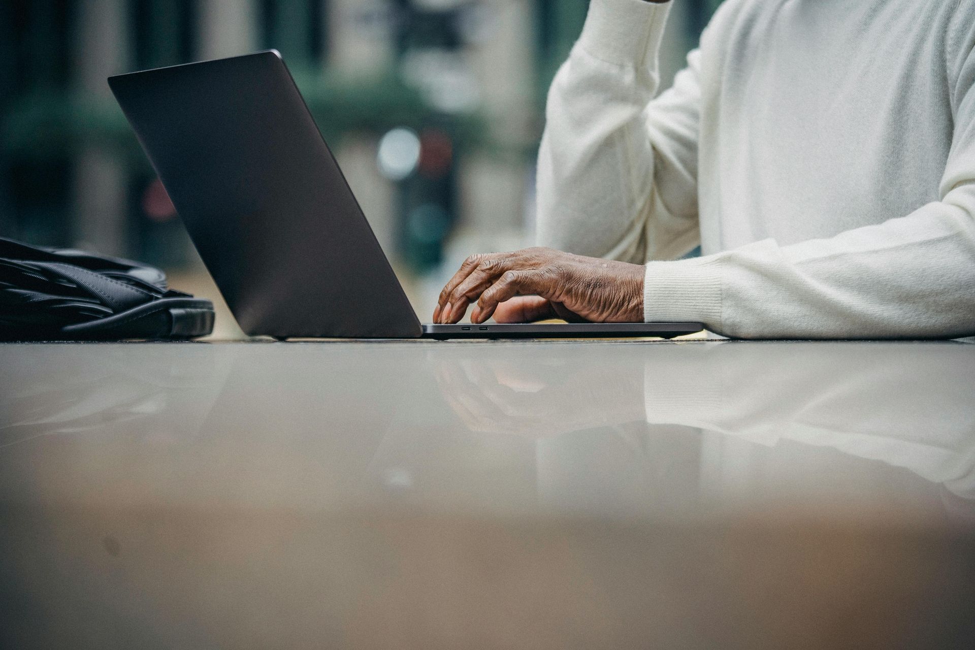 Person typing on a laptop, wearing a white sweater. Laptop sits on a reflective table.