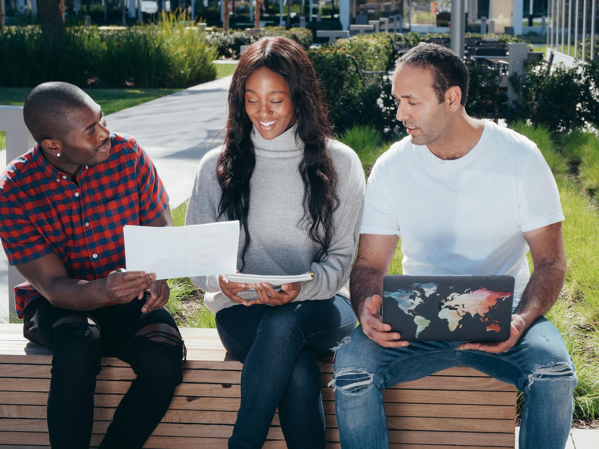 Three diverse people review papers and use a laptop outdoors.