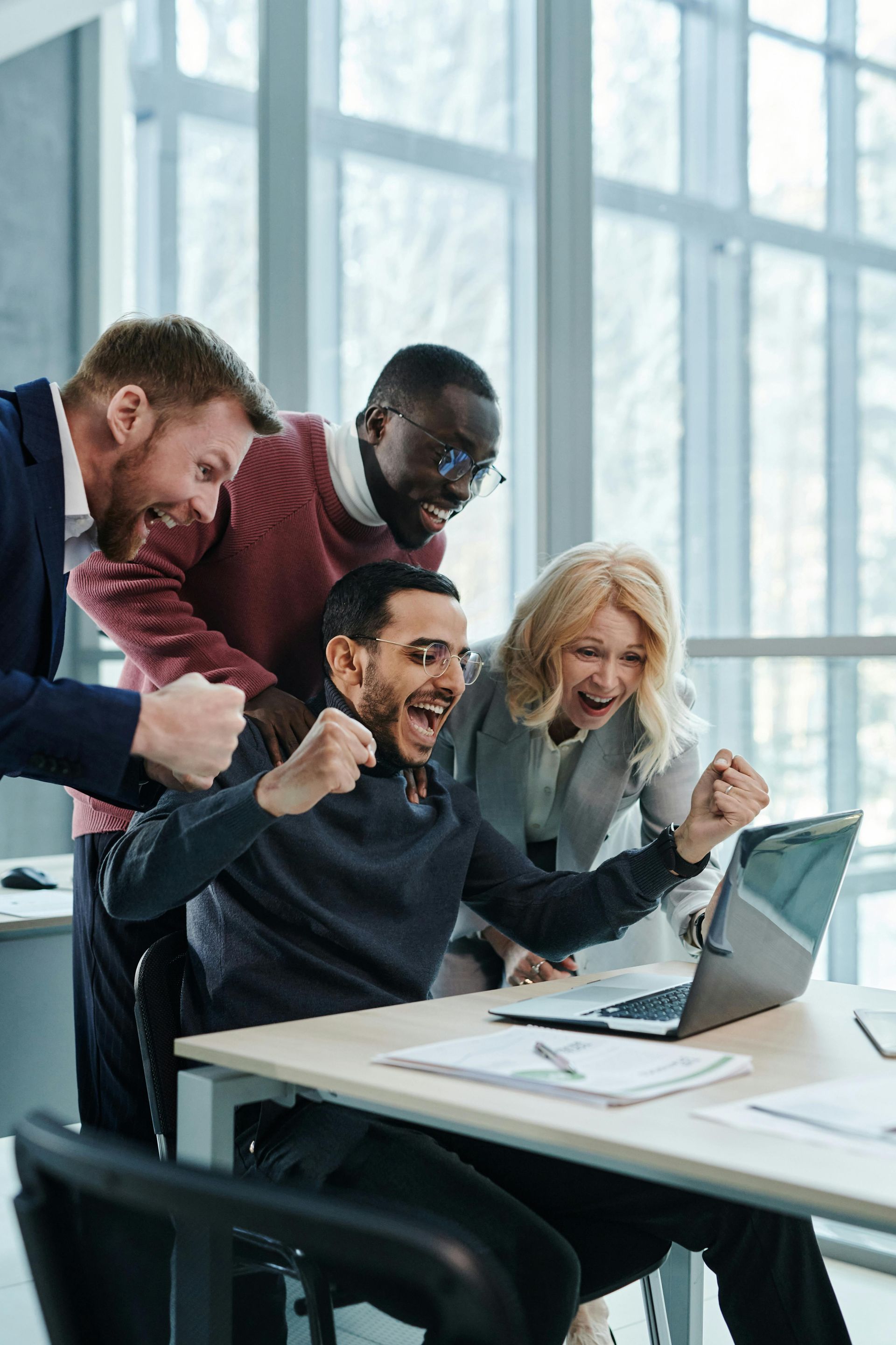 Four excited colleagues celebrate a win while looking at a laptop. Office setting with large windows.