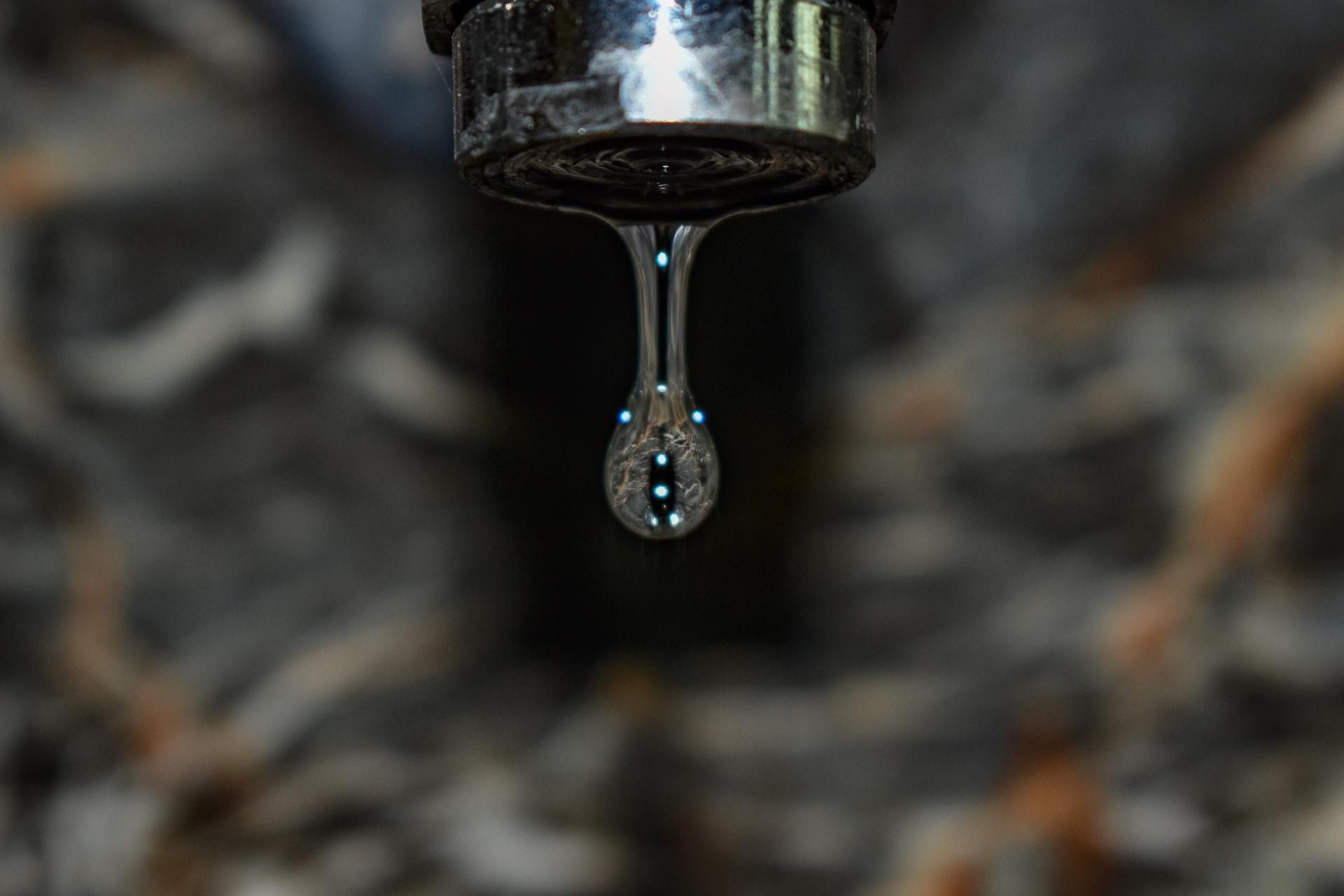 Water droplet hanging from a metal faucet against a blurred dark background