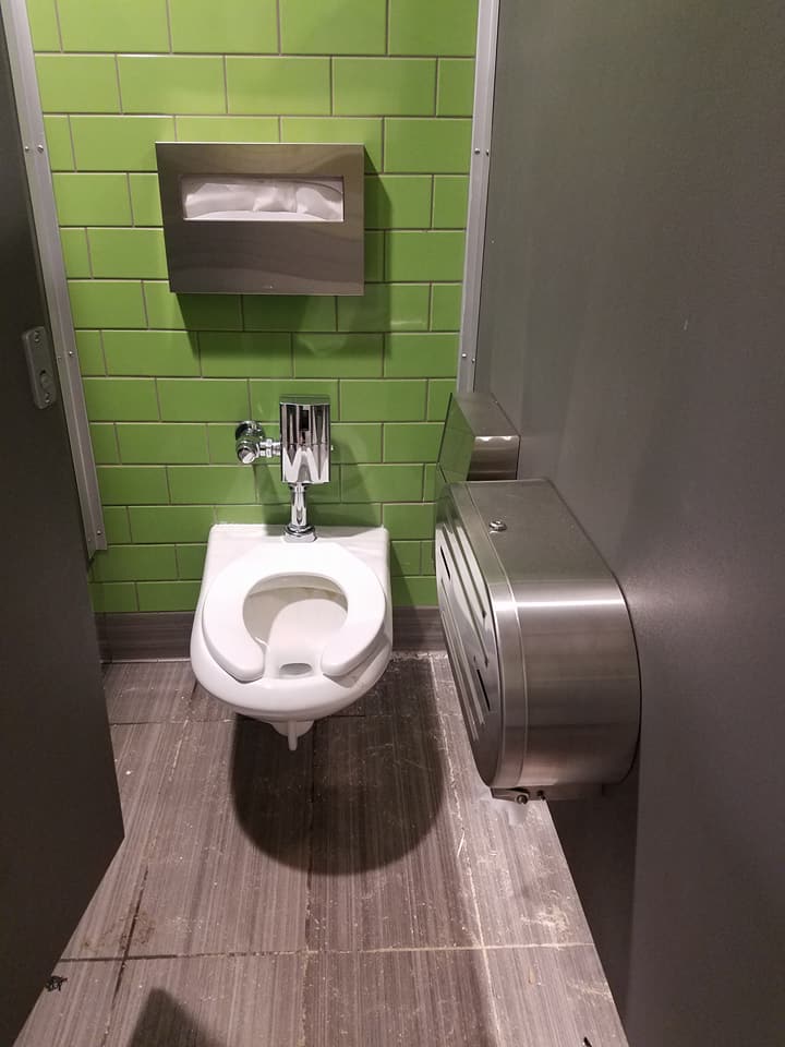 A toilet in a public restroom with green tiles and a paper towel dispenser.