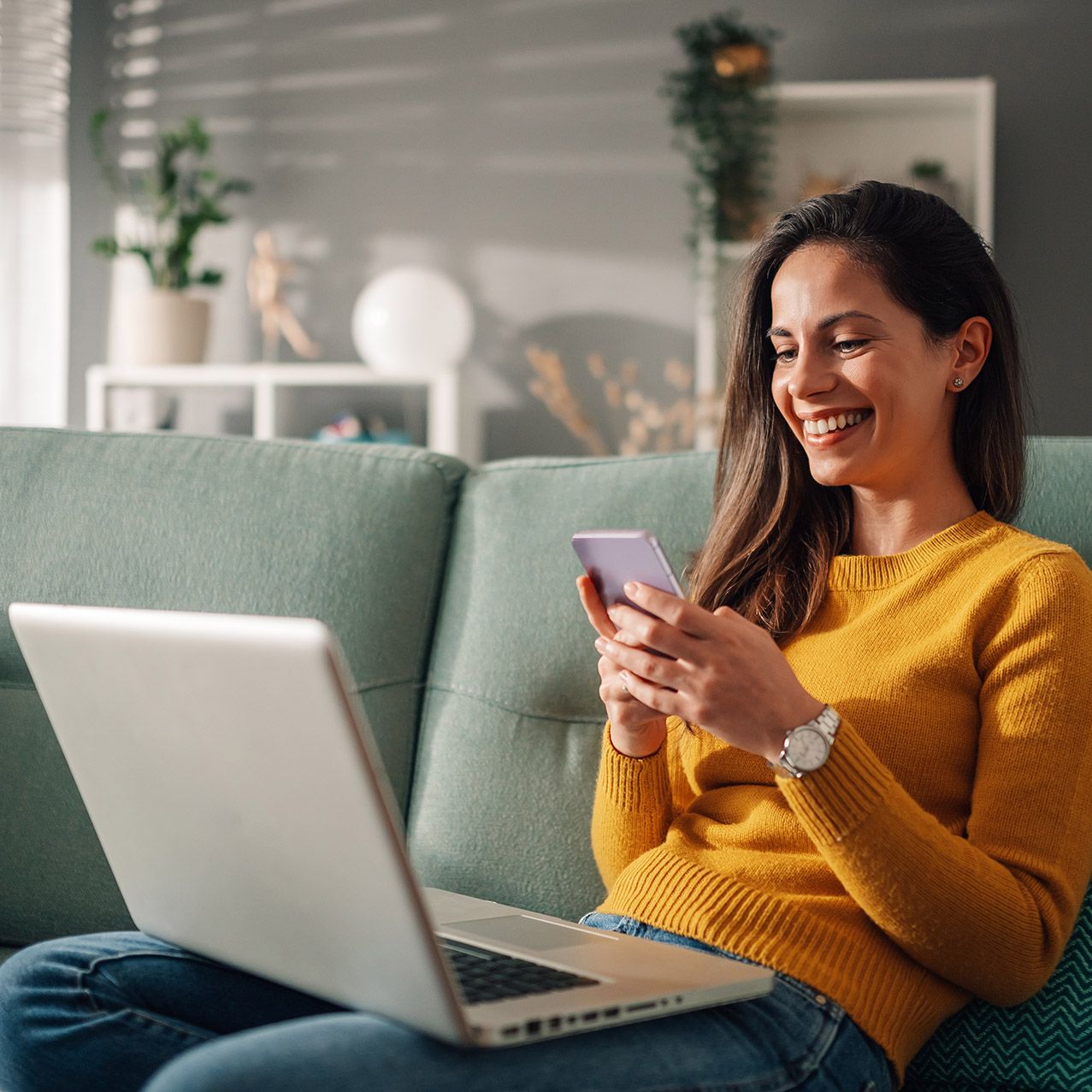 A woman is sitting on a couch using a laptop and a cell phone.