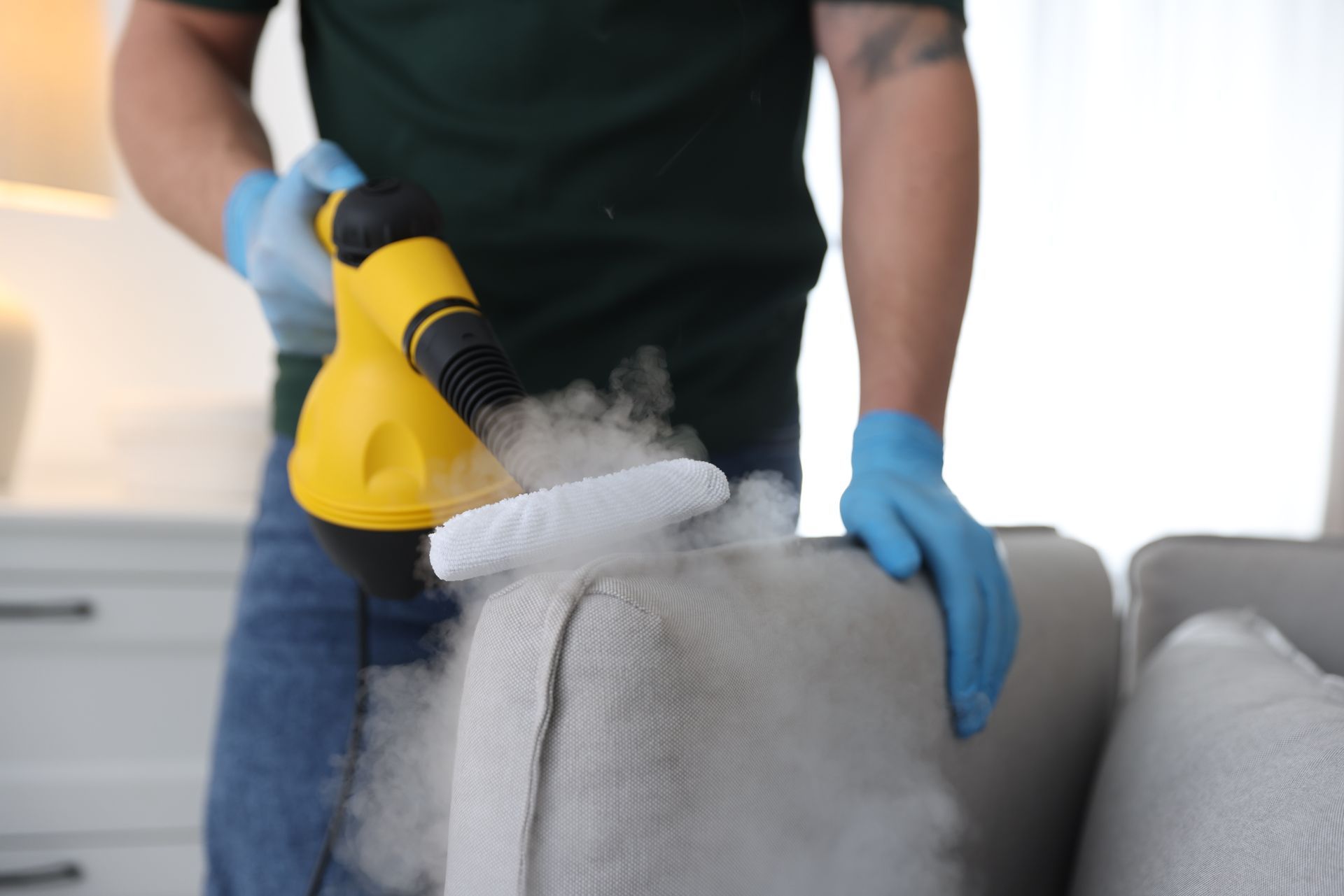 Person steaming a gray sofa with a yellow steam cleaner, wearing blue gloves.
