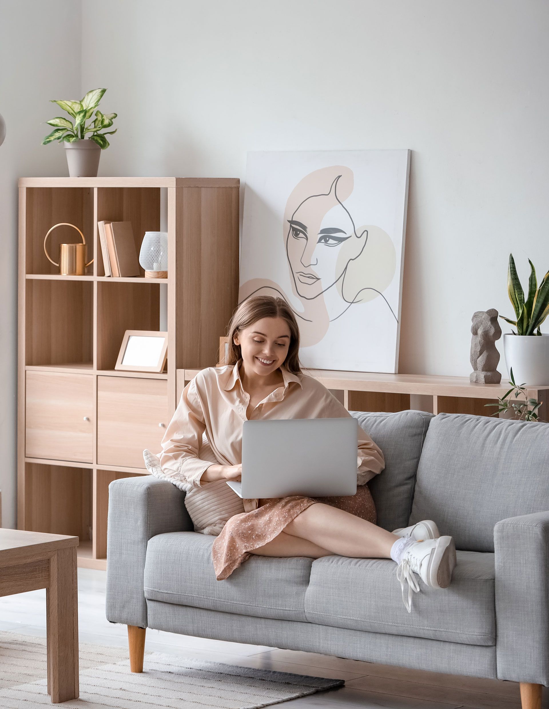Woman using laptop, sitting on gray sofa in light-filled living room.