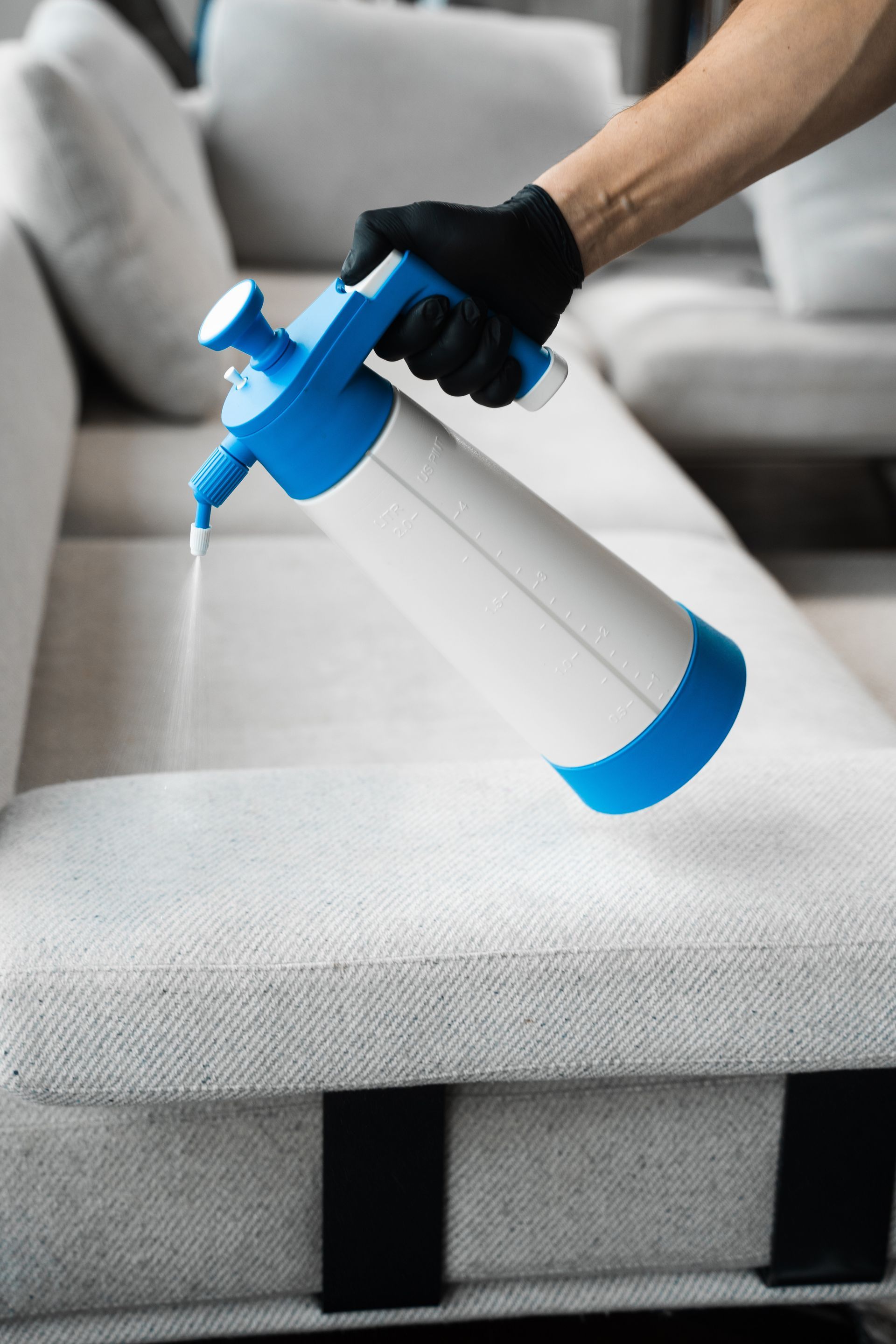 A hand in a black glove spraying a white and blue bottle onto a gray couch.