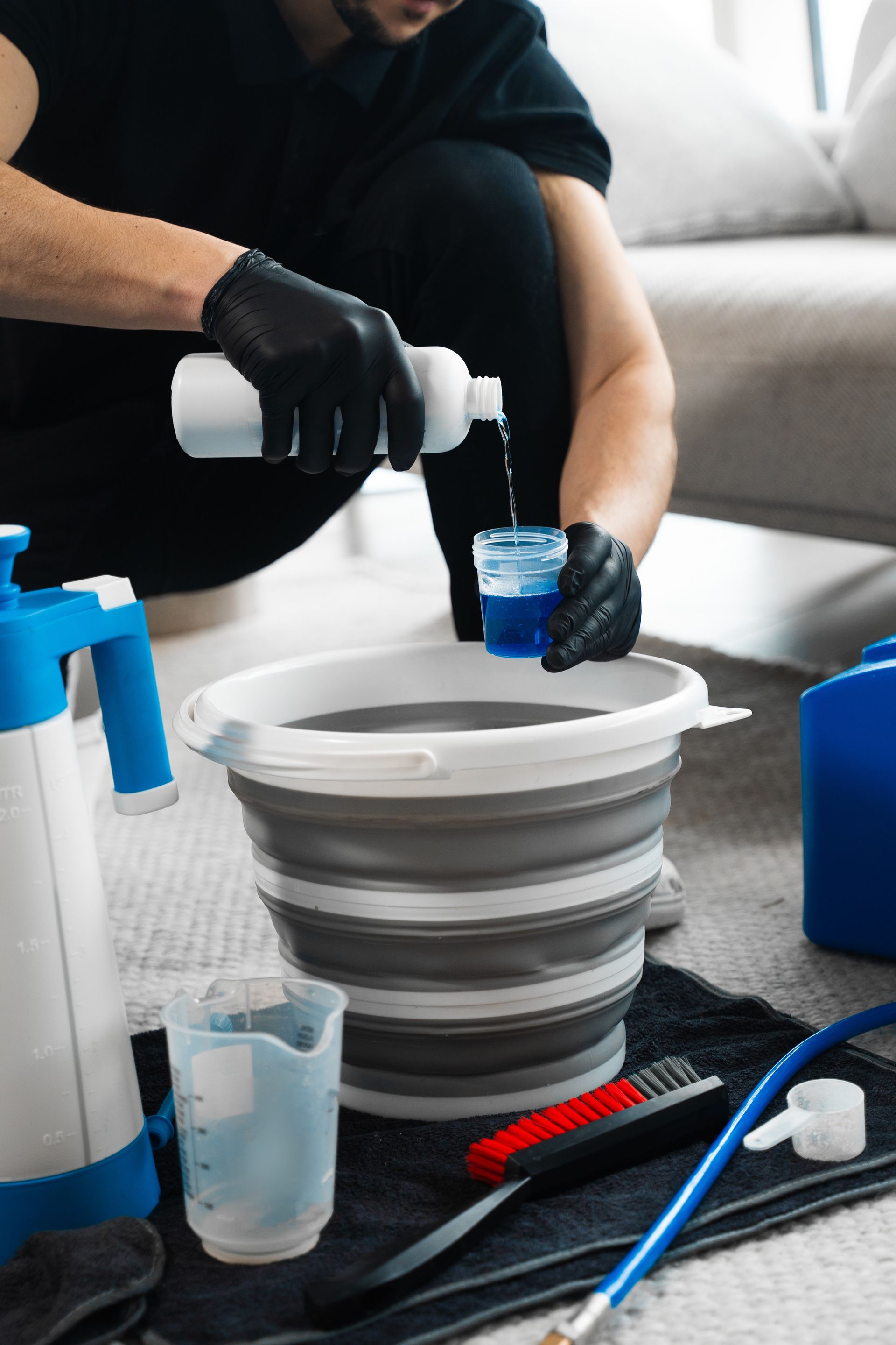 Person pouring blue liquid into a bucket while wearing gloves, preparing to clean a carpet.