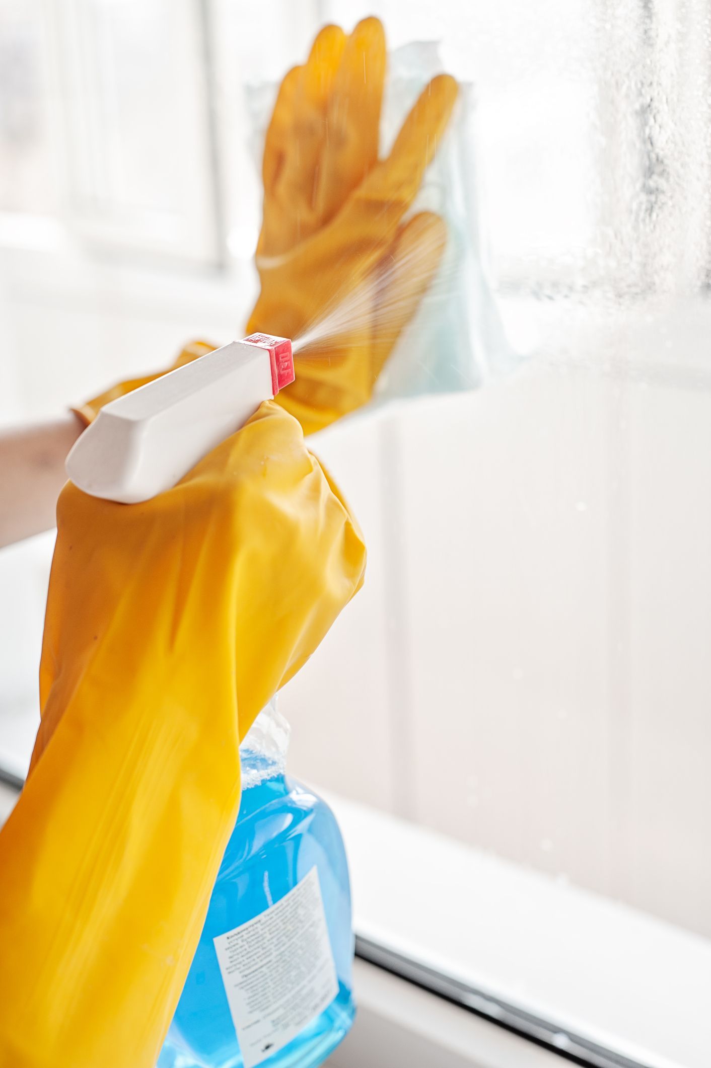 Hands in yellow gloves spraying window cleaner onto a window; wiping with a cloth.