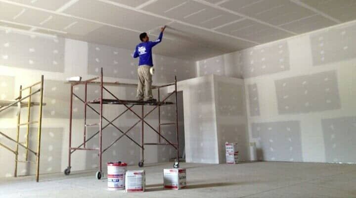 Un trabajador se encuentra sobre un andamio metálico, aplicando masilla para juntas a un techo de paneles de yeso en una habitación sin terminar.