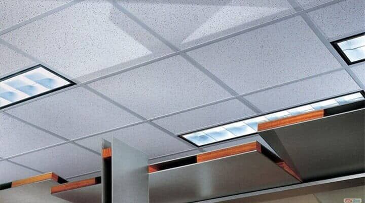 Un falso techo con baldosas blancas cuadradas, luminarias empotradas rectangulares y un elemento arquitectónico moderno en capas.