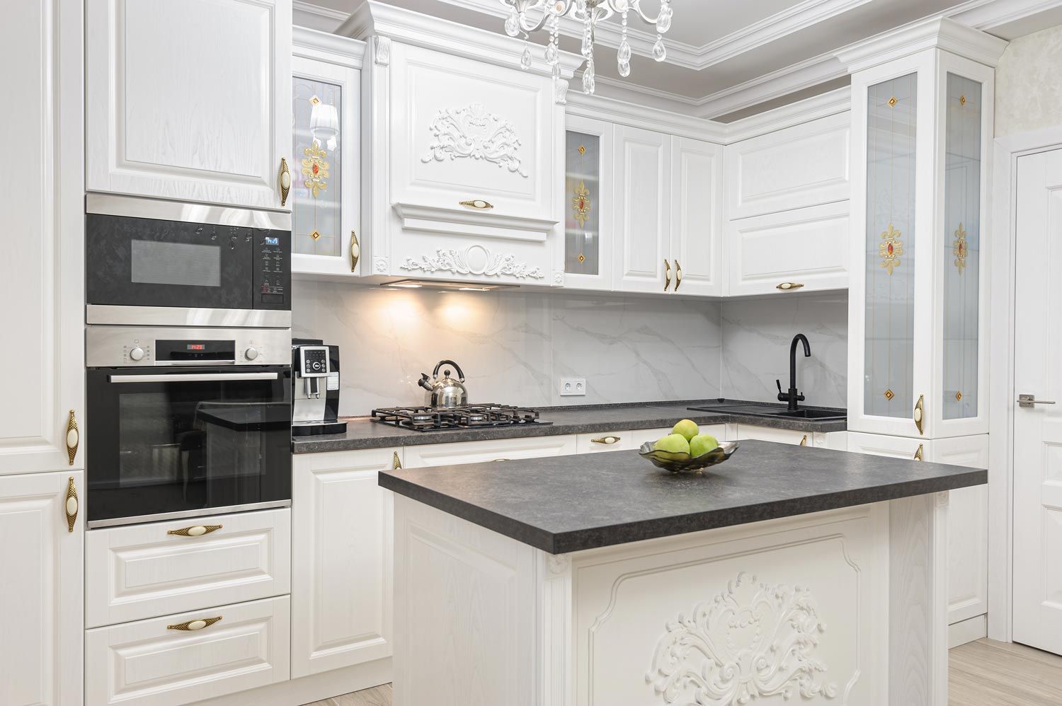 Elegant white kitchen with black countertops, island, built-in oven, and bowl of green apples