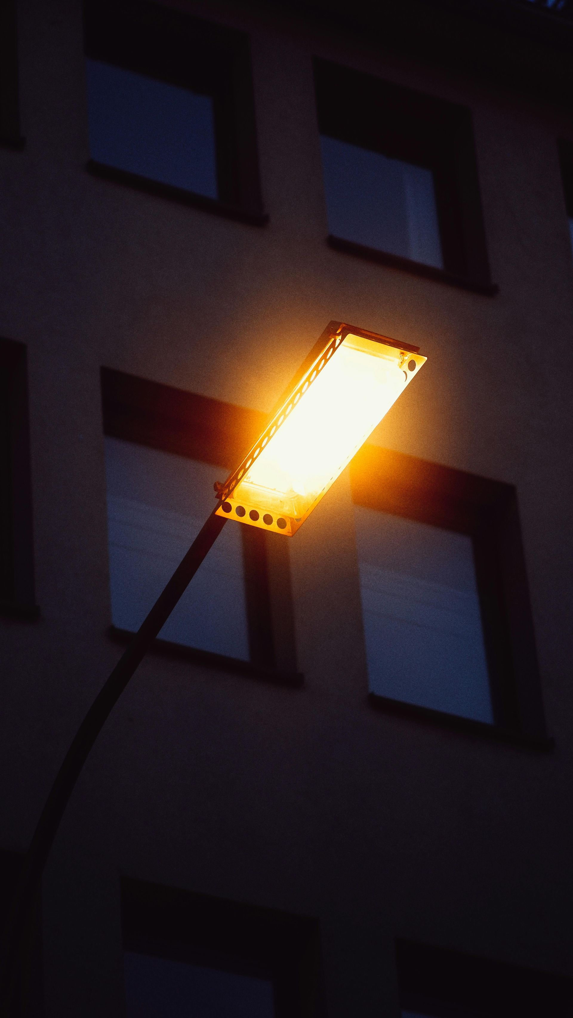 Street lamp illuminates a building with square windows; the lamp is bright orange against the night sky.