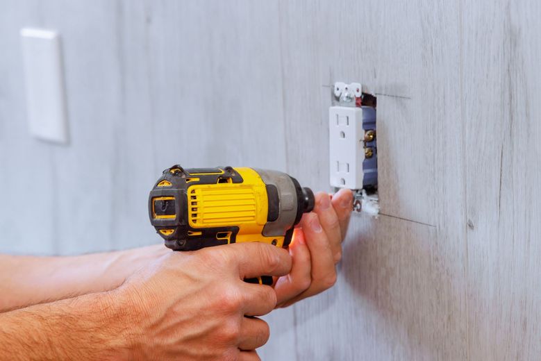 Person using a yellow drill to install an electrical outlet in a wall.