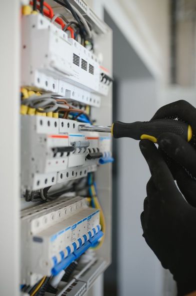Electrician working on a circuit breaker panel with a screwdriver, wearing black gloves.
