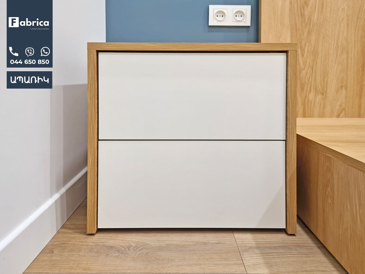 A white nightstand with a wooden frame is next to a bed