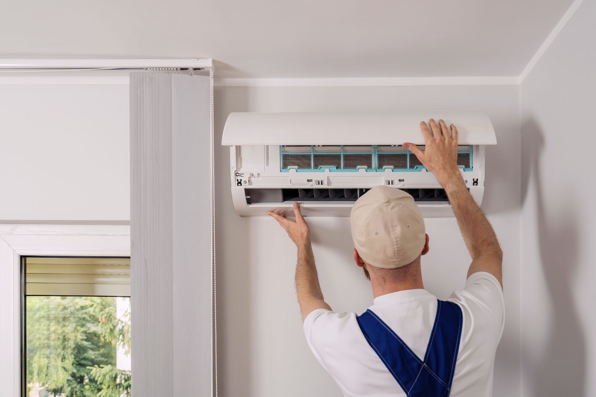 A person in overalls opening an air conditioner unit on a wall near a window.