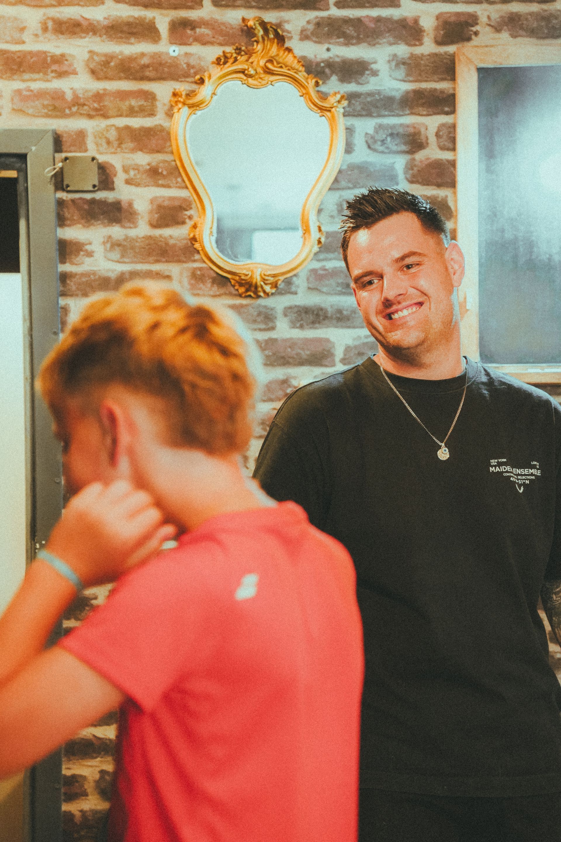 Un homme sourit à la caméra tandis qu'une personne en chemise rouge se cache le visage. Miroir sur un mur de briques.