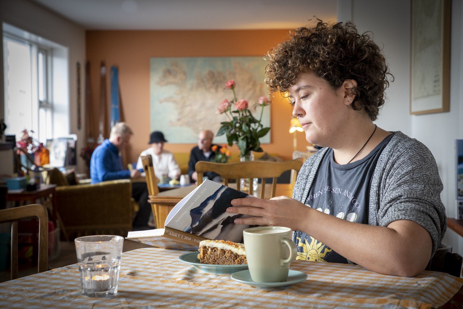 A woman is sitting at a table reading a book and drinking coffee.
