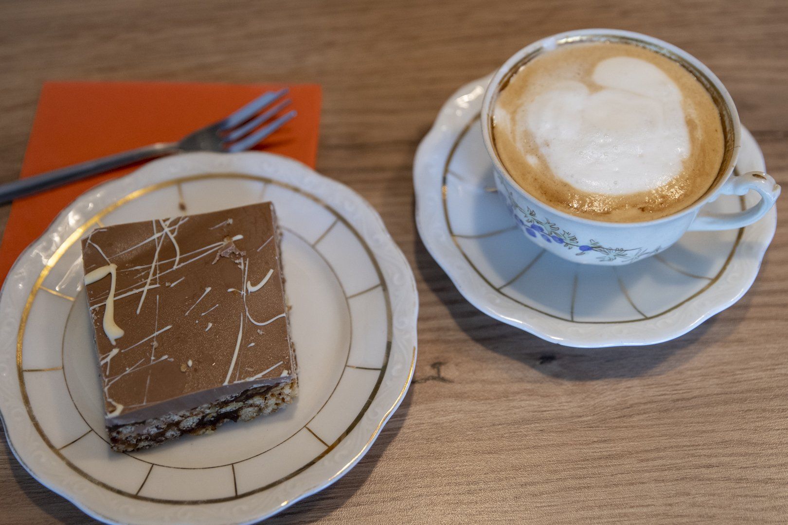 A piece of chocolate cake sits next to a cup of cappuccino