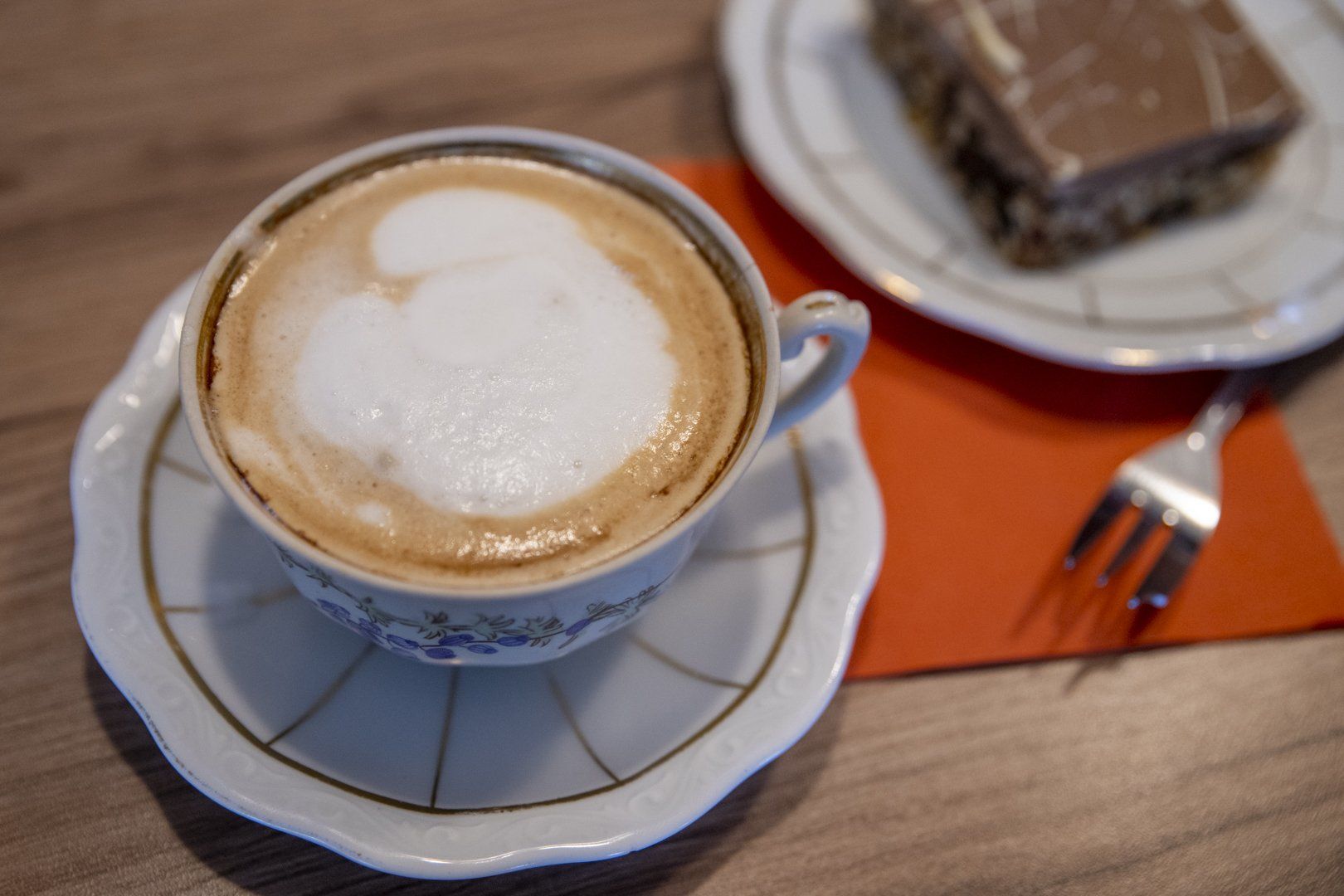 A cup of cappuccino is on a saucer next to a piece of cake.