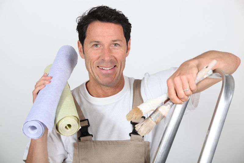 Un homme tient des rouleaux de papier peint et des pinceaux tout en s'appuyant sur une échelle.