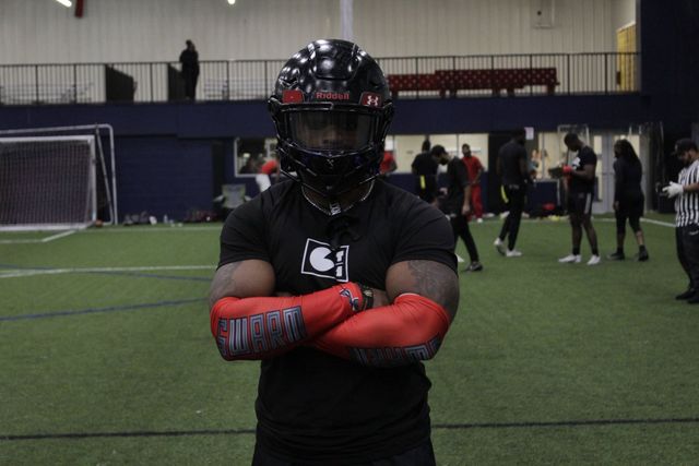Football player in helmet and pads with arms crossed on indoor field.