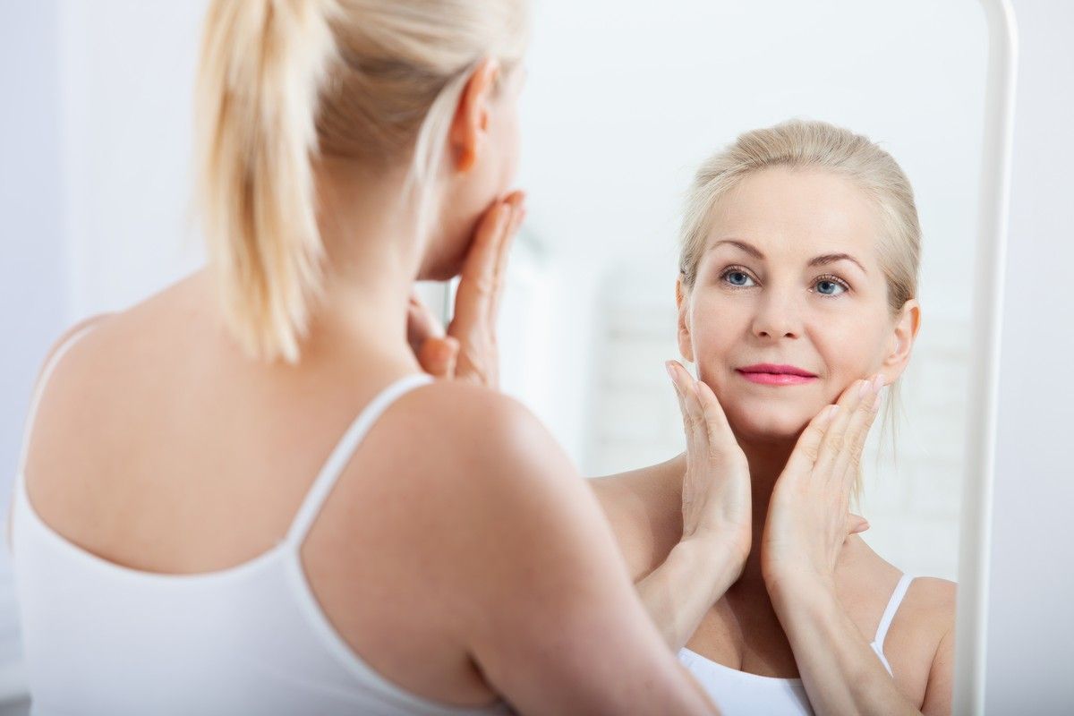 Woman touching face in mirror, representing anti-aging facials to reduce fine lines and firm skin in Albuquerque.