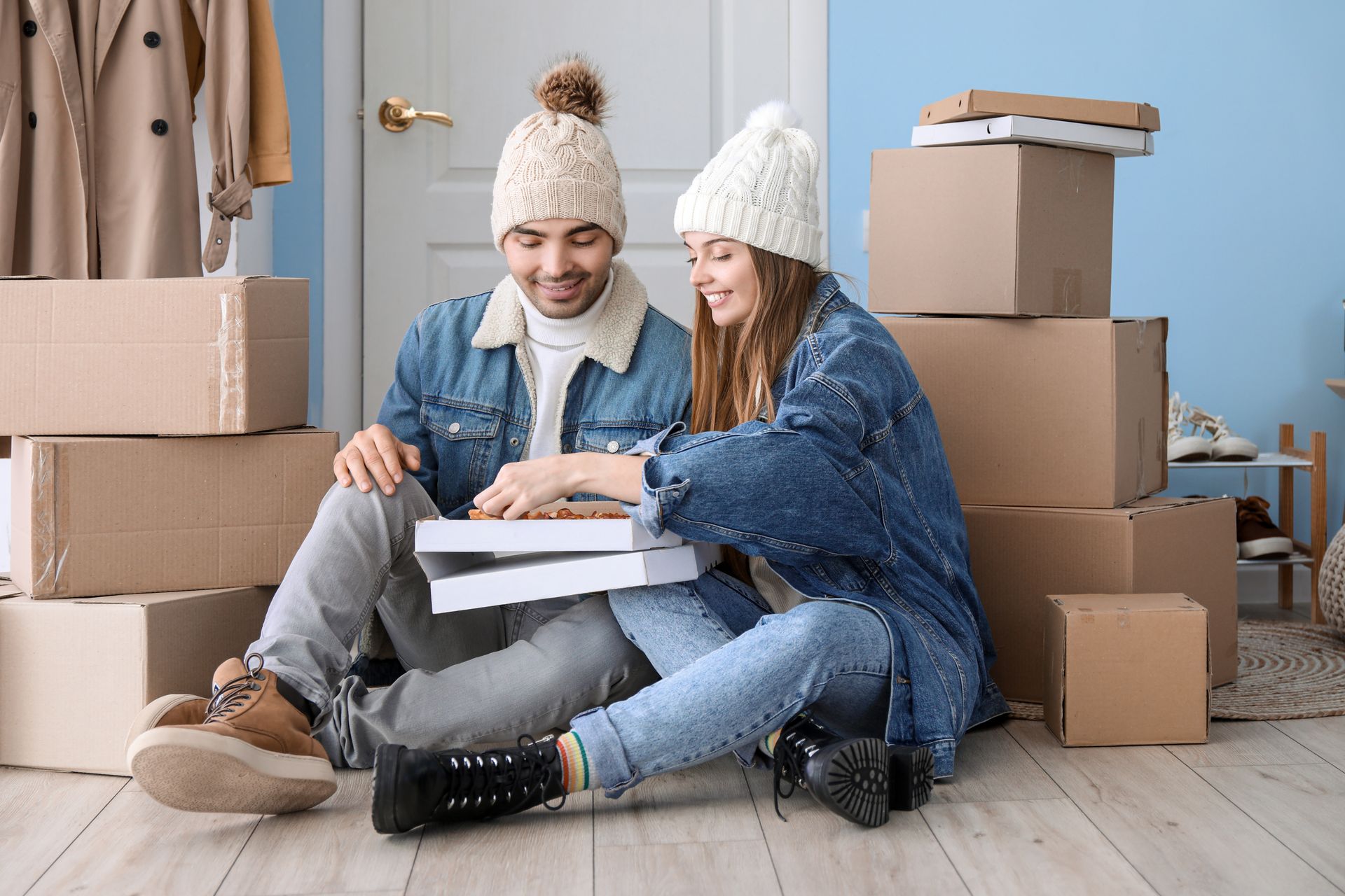 Couple sits on the floor surrounded by boxes, eating from a pizza box, both wearing winter hats.