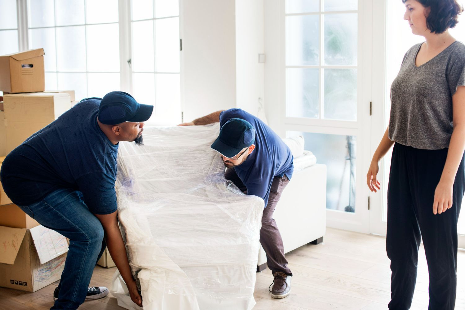 Two movers in blue uniforms wrap a white sofa in plastic inside a room while a person stands nearby watching.