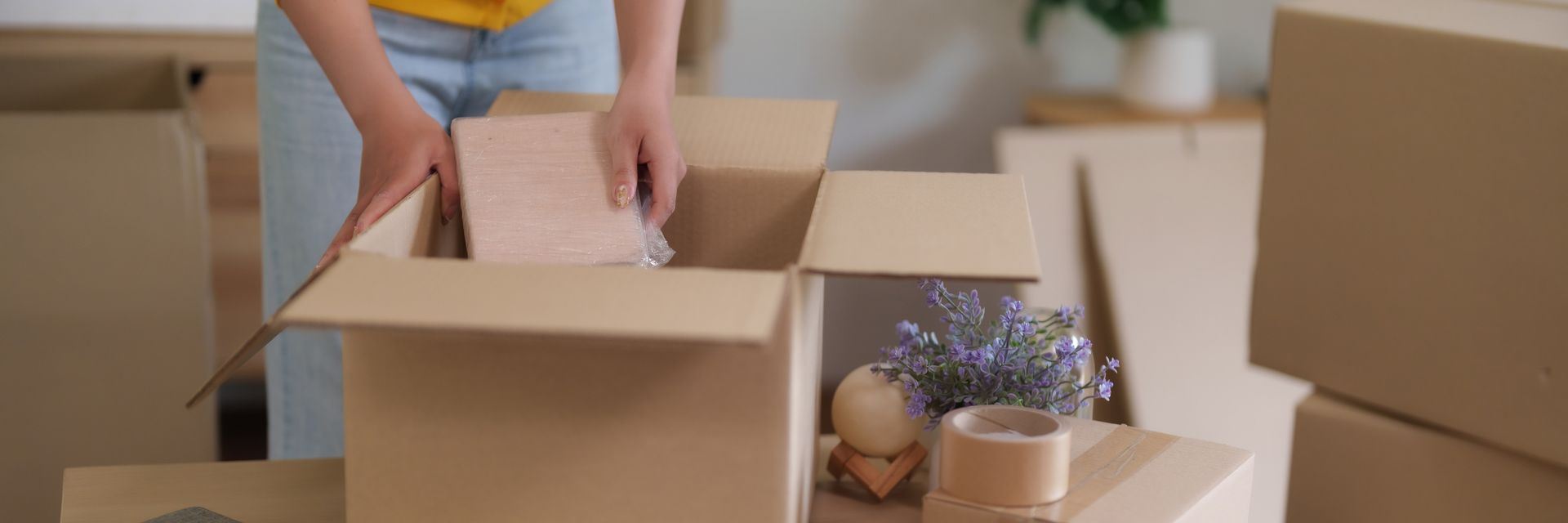Person packing a cardboard box, likely for moving. Hands are placing items into the box, other boxes are nearby.