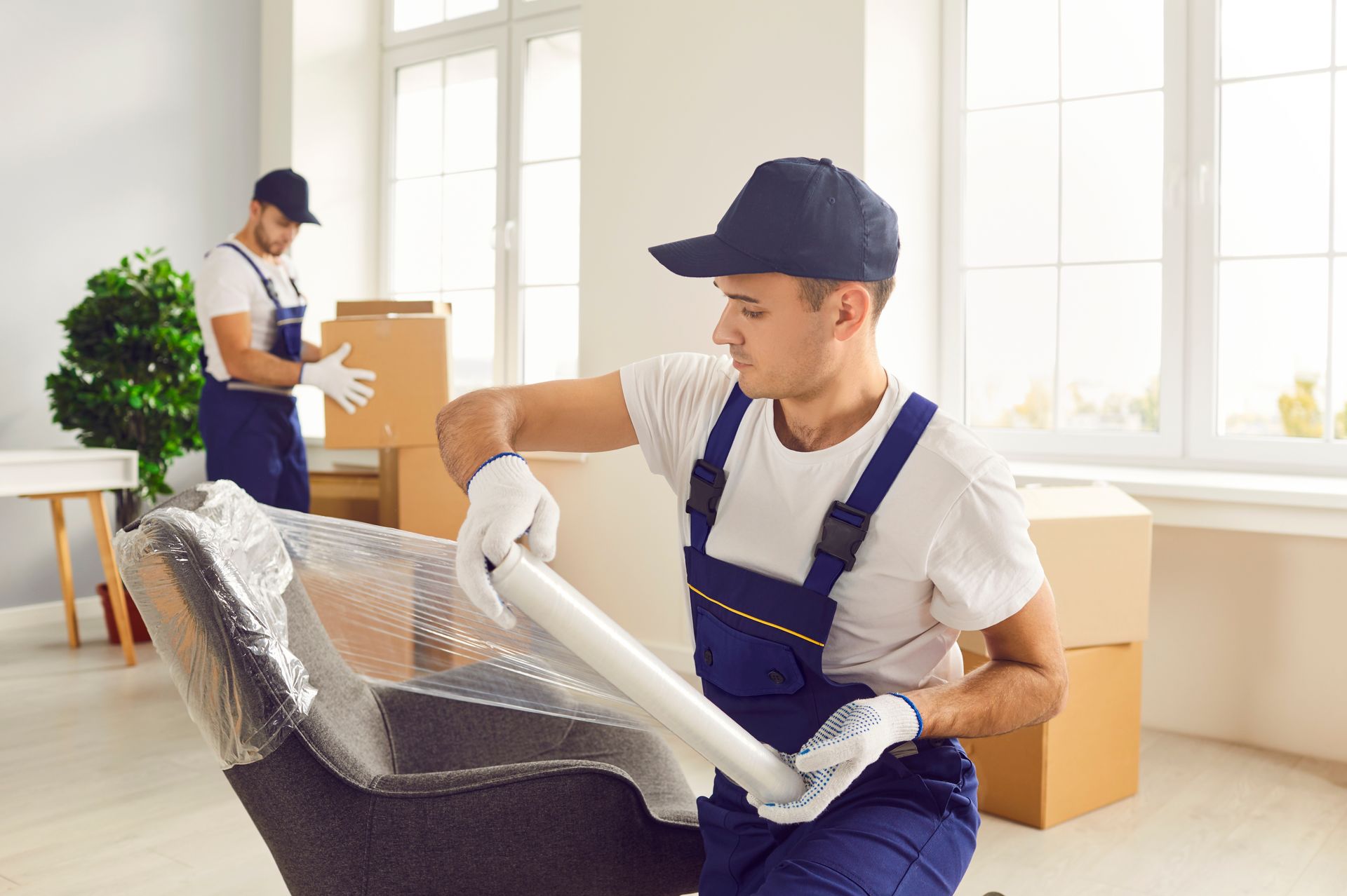 Two movers wrapping furniture with plastic in a brightly lit room with boxes.
