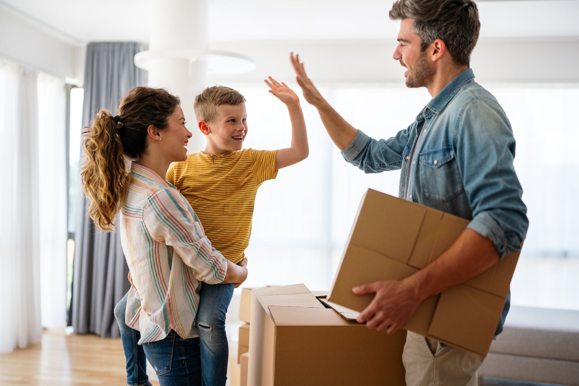 Family high-fives in new home holding boxes; mom, dad and son celebrating moving day.