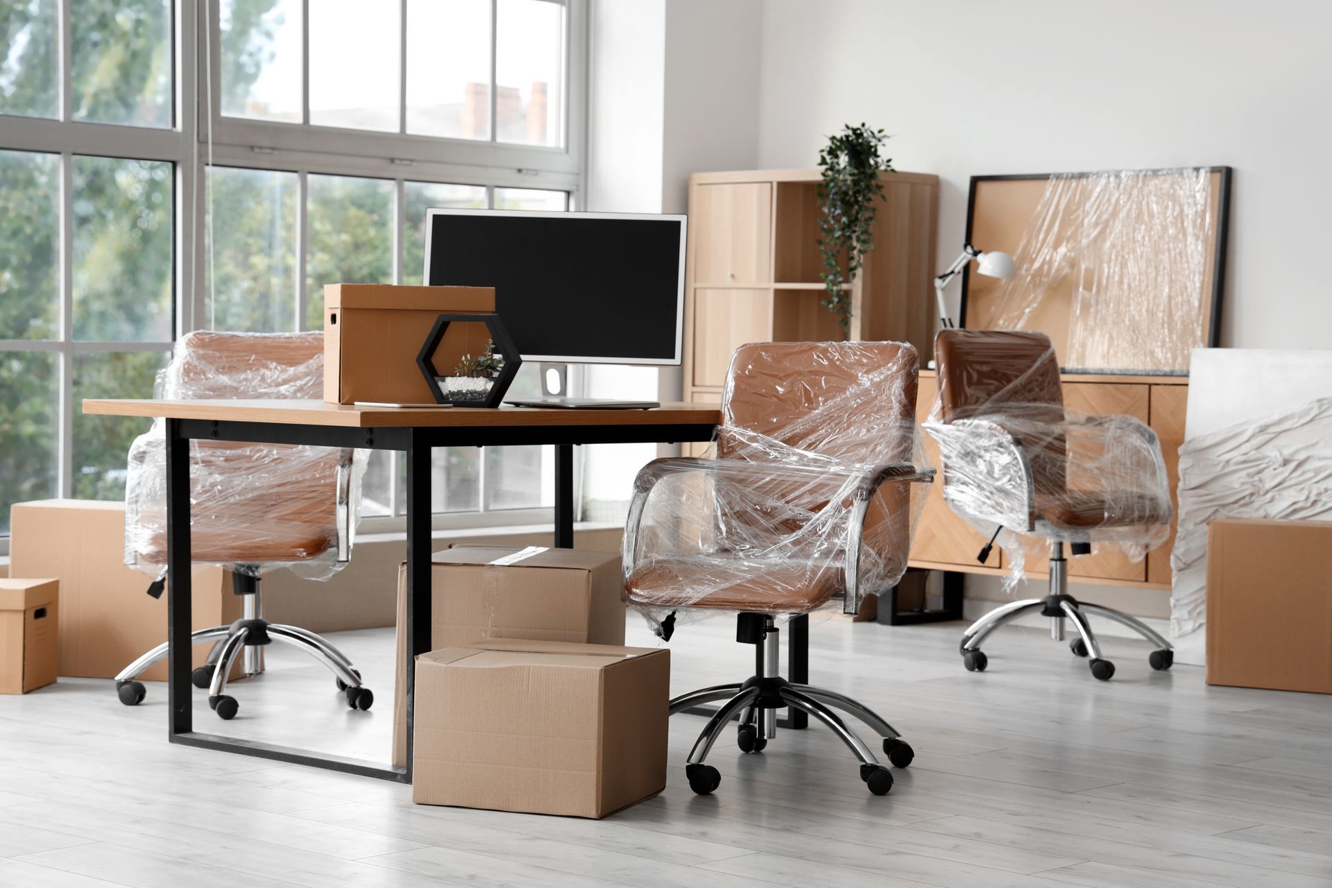 Office being moved, furniture wrapped in plastic, cardboard boxes. Window in the background.