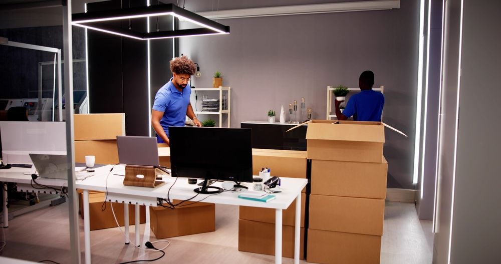 Two people in blue shirts organize an office space filled with stacked cardboard moving boxes and desk equipment.