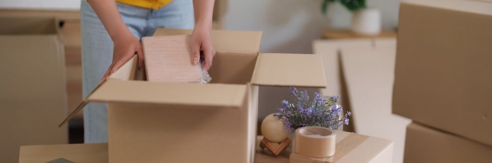 A person packing a light-colored object into an open cardboard moving box in a room with other boxes.