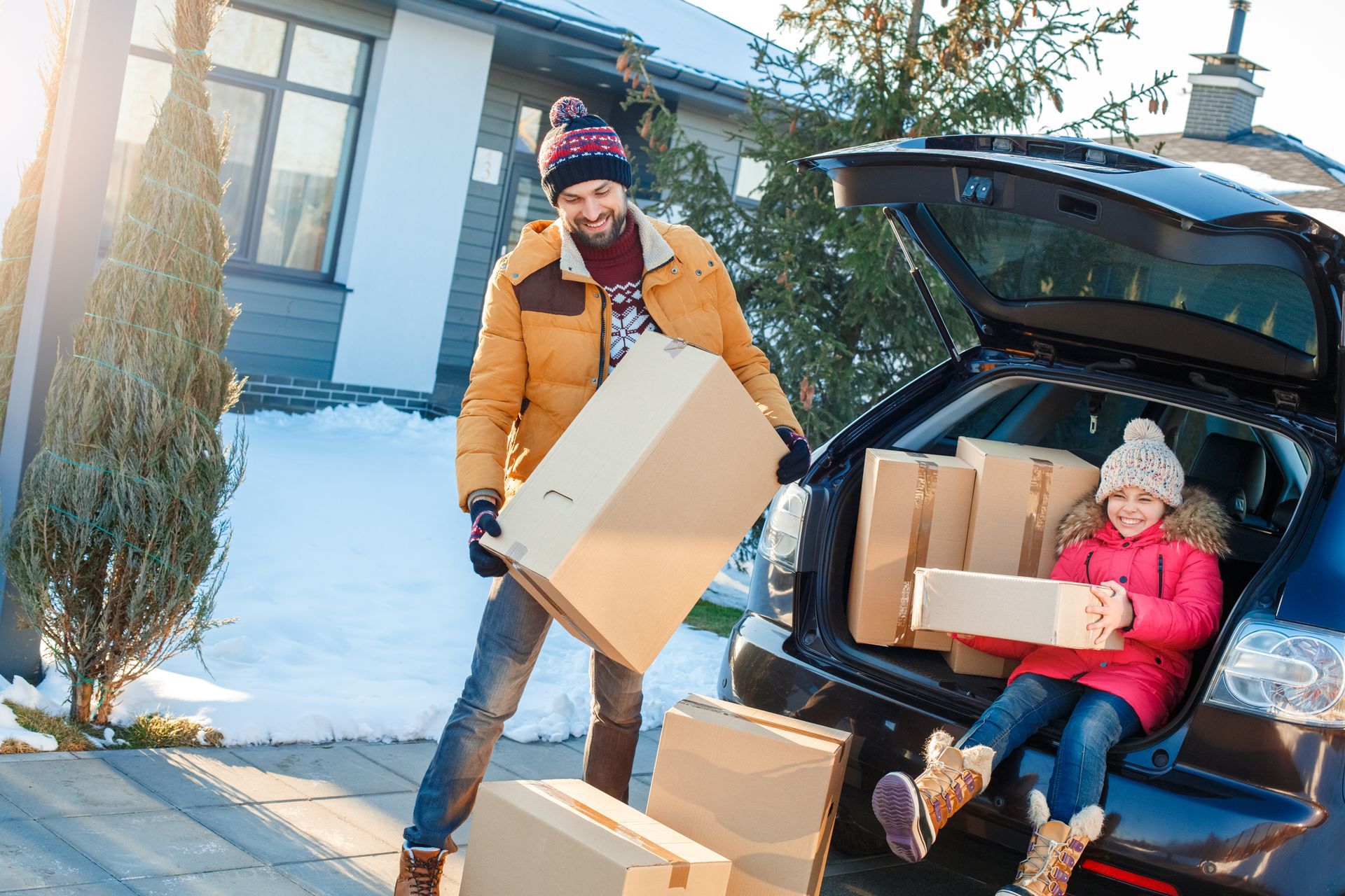 Father and daughter load boxes into a car trunk in the snow; both are wearing winter clothing.
