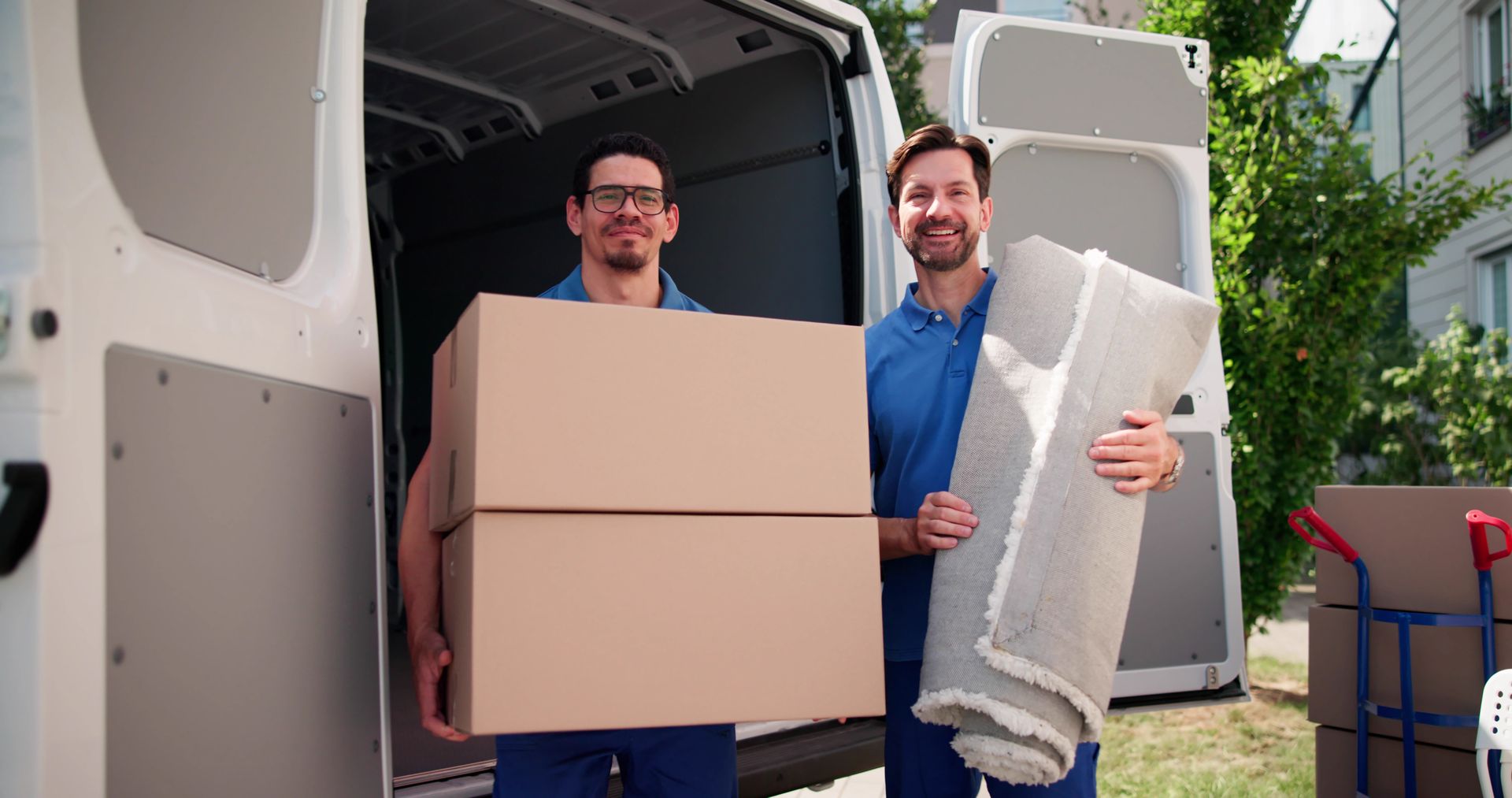 Two movers smiling, holding boxes and a rolled rug next to an open van.