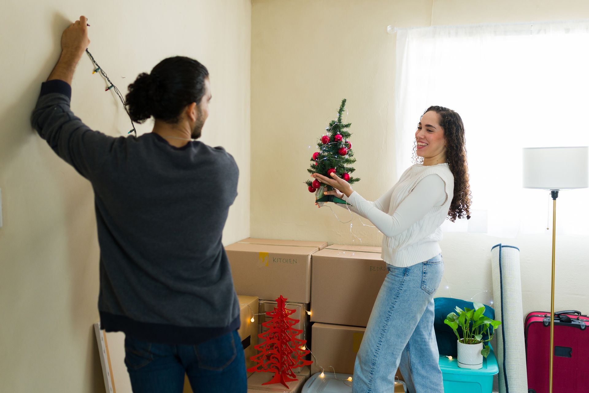Couple decorating a new home; man hangs lights, woman holds small Christmas tree, smiles. Boxes in the background.