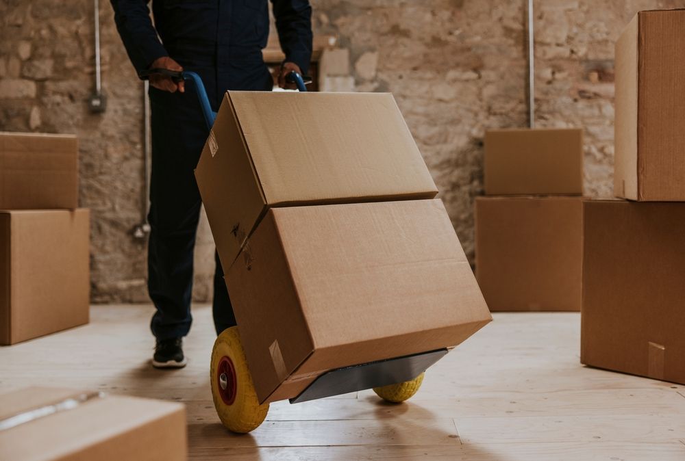A person in uniform uses a yellow-wheeled hand truck to transport two stacked cardboard boxes in a storage room.