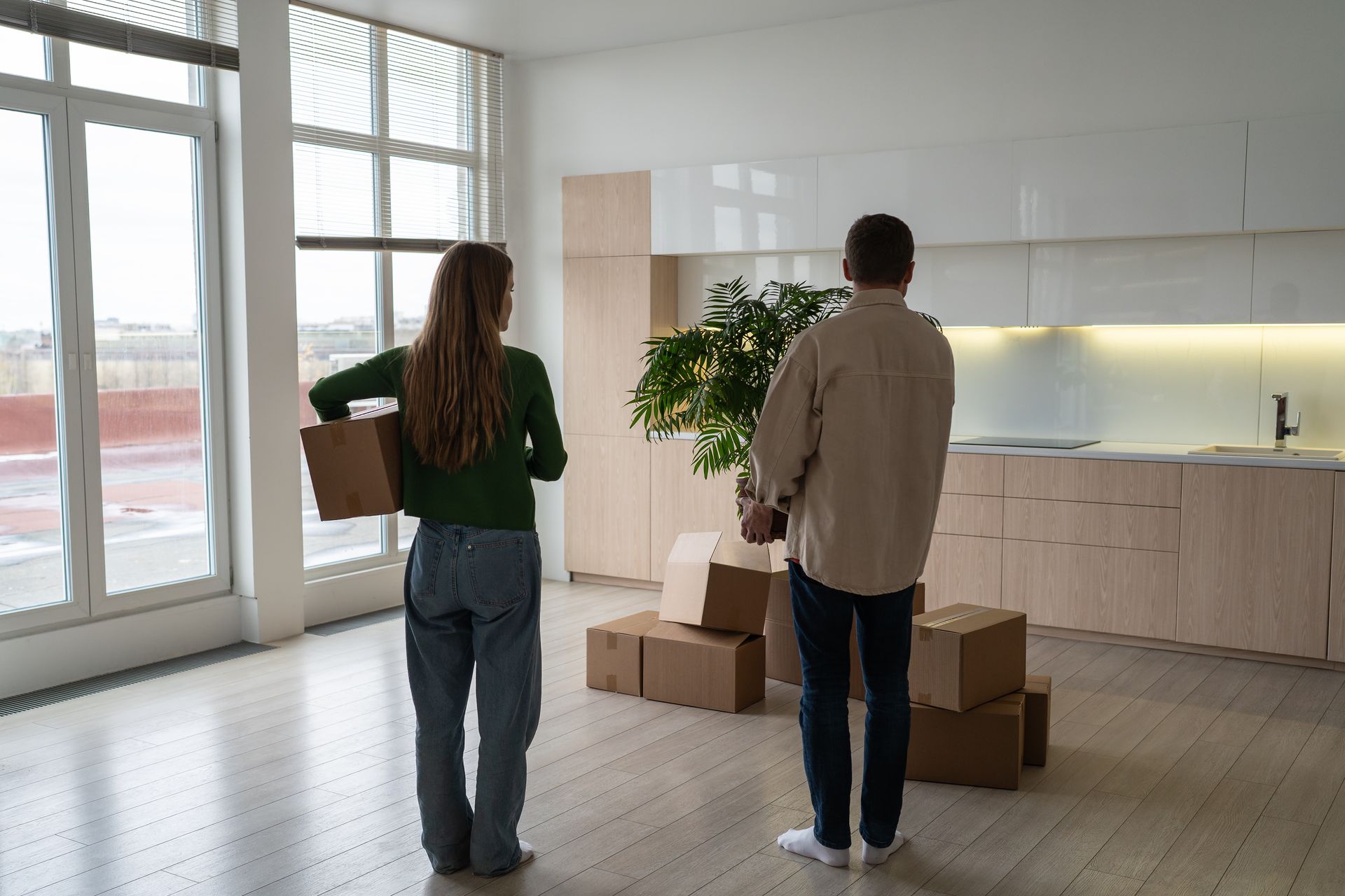 Couple in new, empty apartment, holding boxes, near stacked boxes and kitchen cabinets.