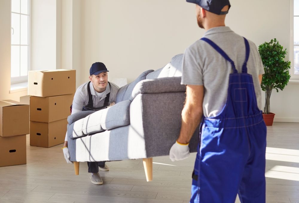 Two movers in uniforms and gloves carry a gray sofa into an empty, sunlit room with cardboard moving boxes.